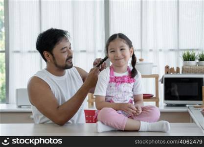 Happy Asian young father with beard making braid for his Caucasian little daughter while sitting on table at home kitchen. Single dad try to plaiting cute girls hair in the morning weekend with love