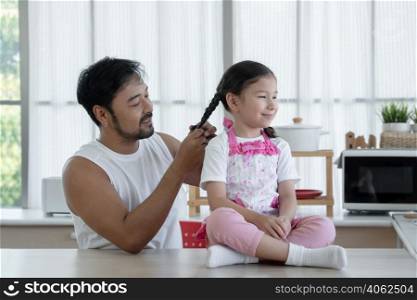 Happy Asian young father with beard making braid for his Caucasian little daughter while sitting on table at home kitchen. Single dad try to plaiting cute girls hair in the morning weekend with love