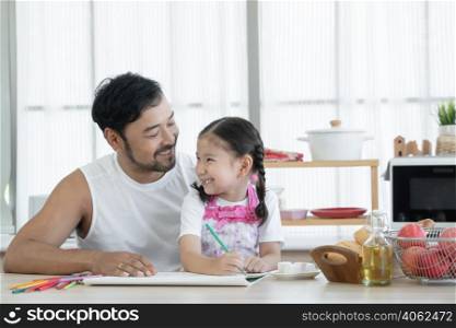 Happy Asian young father with beard smiling at his cute daughter while drawing with colored pencil together at kitchen at home with marshmallows, breads and red apples on table. Children education