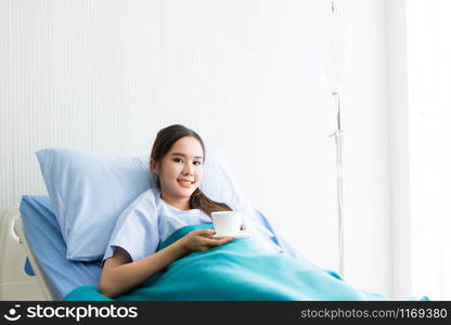 Happy asian young female patient on bed with hand holding a cup coffee or tea in hospital background