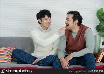 Happy Asian young gay couple wearing sweater feeding his boyfriend strawberry and sitting on sofa together at living room at home. Romance Lgbt family in Valentine day