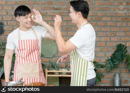 Happy Asian young LGBT gay couple cooking together. Handsome men playing red tomato and cucumber sliced and put on eye in kitchen at home. Relationship of homosexual lifestyle concept