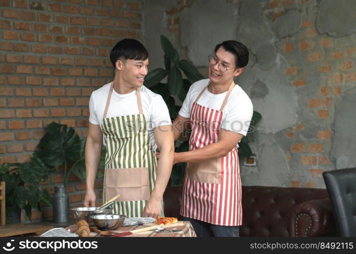 Happy Asian young LGBT gay couple making pizza together. A man helping his boyfriend put on an apron in kitchen at home. Relationship of homosexual lifestyle concept
