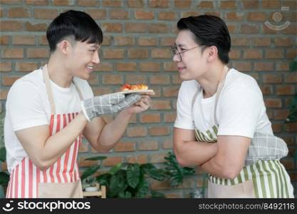Happy Asian young LGBT gay couple making pizza together. Handsome man showing sliced homemade pizza to his boyfriend for smelling in kitchen at home. Relationship of homosexual lifestyle concept