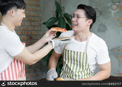 Happy Asian young LGBT gay couple making pizza together. Handsome man feeding sliced homemade pizza to his boyfriend tasting in kitchen at home. Relationship of homosexual lifestyle concept