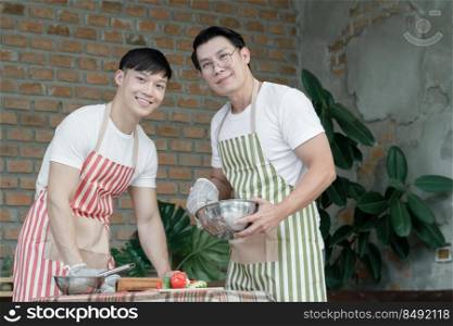 Happy Asian young LGBT gay couple with apron cooking together. Handsome men mixing salad dressing and cutting tomato cucumber in kitchen at home. Relationship of homosexual lifestyle concept