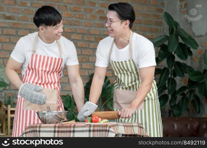 Happy Asian young LGBT gay couple with apron cooking together. Handsome men using rolling pin and cutting tomato cucumber to make salad in kitchen at home. Relationship of homosexual lifestyle concept