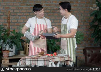 Happy Asian young LGBT gay couple with apron cooking together. Handsome man mixing salad dressing and his boyfriend thumb up to him and using tablet in kitchen at home.