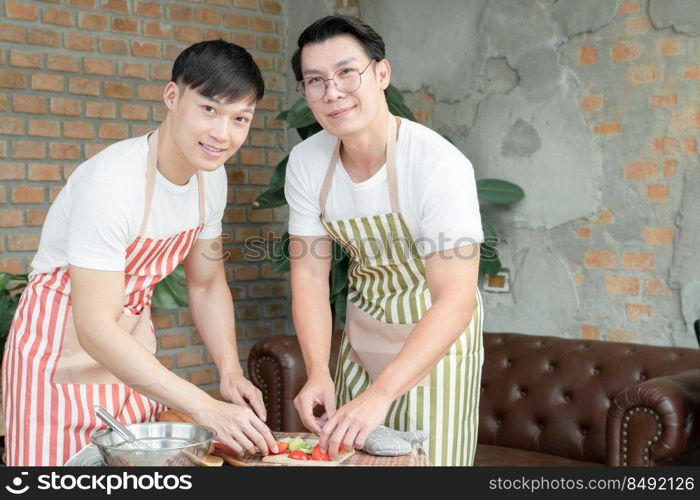 Happy Asian young LGBT gay couple with apron cooking together. Handsome men cutting tomato cucumber to make salad in kitchen at home. Relationship of homosexual lifestyle concept