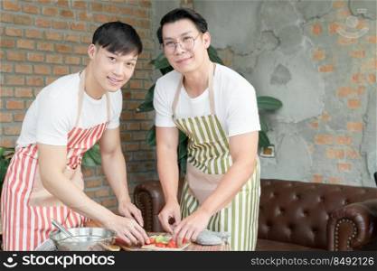 Happy Asian young LGBT gay couple with apron cooking together. Handsome men cutting tomato cucumber to make salad in kitchen at home. Relationship of homosexual lifestyle concept