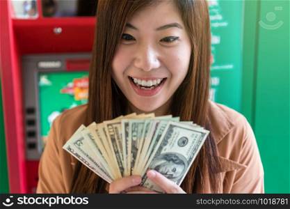 Happy asian young woman holding the dollar money with smiling action after withdrawed cash at an ATM, business automatic money transfer concept