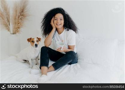 Happy attractive ethnic woman with curly hair wears white t shirt, jeans and socks, smiles pleasantly, drinks tea in comfortable bed, poses with dog, have lazy weekend. People, rest, animals concept
