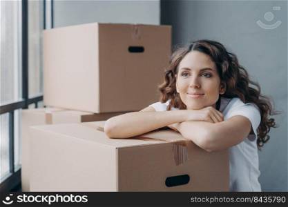 Happy attractive european woman unpacking things and being leaned to boxes. Girl is dreaming near window while sitting on floor in new apartment. Concept of future and perspective.. Happy european woman unpacking things leaned to boxes. Girl dreaming near window in new apartment.