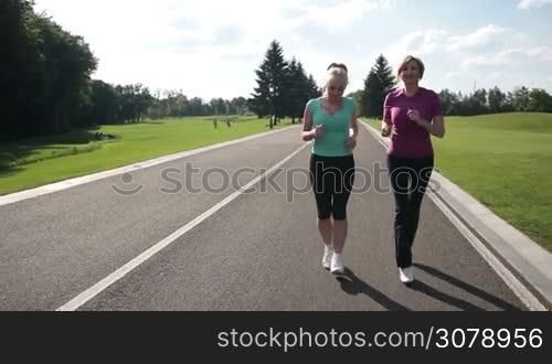 Happy attractive senior females in sport clothes jogging in the park along the road. Healthy fitness women running on park road in sportswear outdoors. Slow motion. Steadicam stabilized shot.