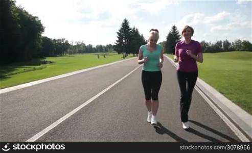 Happy attractive senior females in sport clothes jogging in the park along the road. Healthy fitness women running on park road in sportswear outdoors. Slow motion. Steadicam stabilized shot.