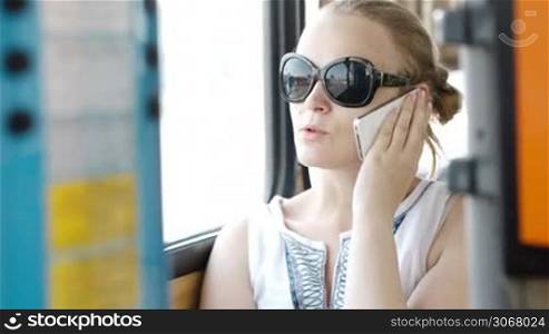 Happy attractive young woman wearing sunglasses talking on her smartphone and smiling with delight while travelling by bus