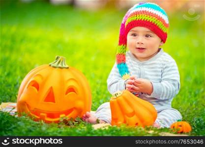 Happy baby boy on Halloween holiday outdoors, sitting on green grass backyard, wearing funny festive colorful hat, playing with cute carved pumpkins