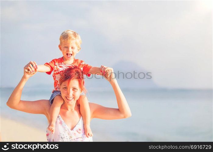 Happy baby boy sitting on shoulders of mother on beach.