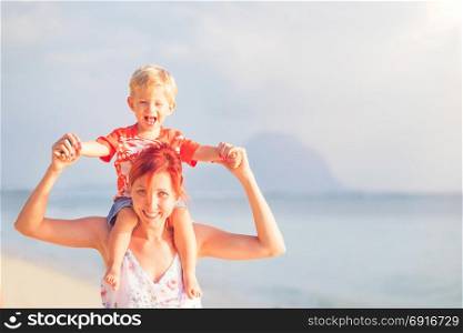 Happy baby boy sitting on shoulders of mother on beach.