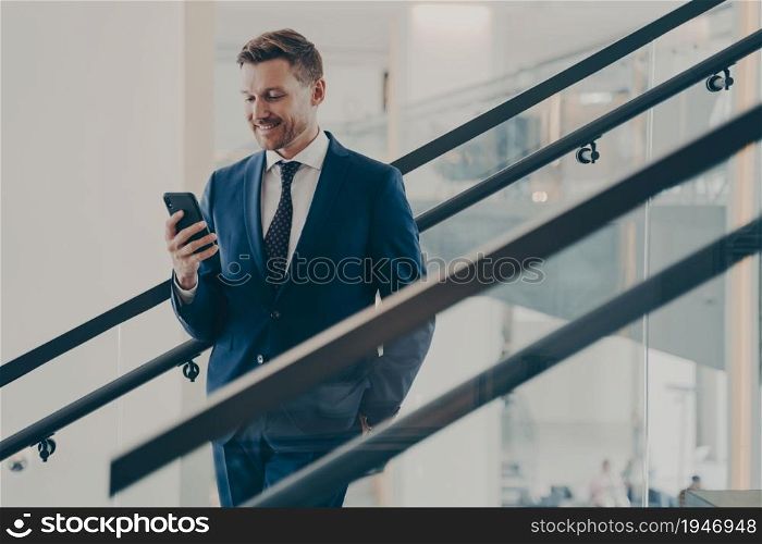 Happy bearded businessman with smartphone standing on staircase. Satisfied young man using mobile phone app, browsing internet, looks at smartphone with smile. Mobile technology and business concept. Businessman in formal suit using smartphone while standing on staircase
