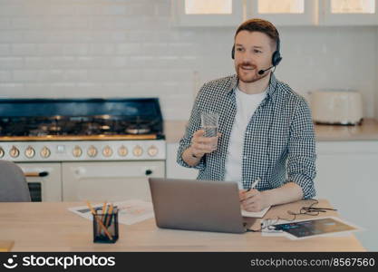 Happy bearded guy in casual clothes and headset working on laptop remotely at home, holding glass of water and making some note while studying online. Remote work and distant education concept. Smiling young man making notes in notebook while studying at home