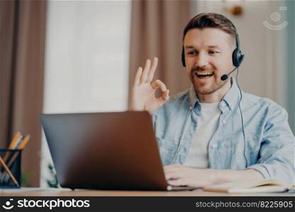Happy bearded guy male freelancer showing ok sign gesturing during online call while sitting at his workplace with laptop in room, man working remotely at home. Freelance and online communication. Smiling stylish man in headset showing ok gesture while having video call