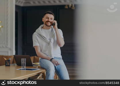 Happy bearded male freelancer works in home office room dressed in casual t shirt and jeans has telephone conversation discusses working issues has cheerful expression works remotely from home