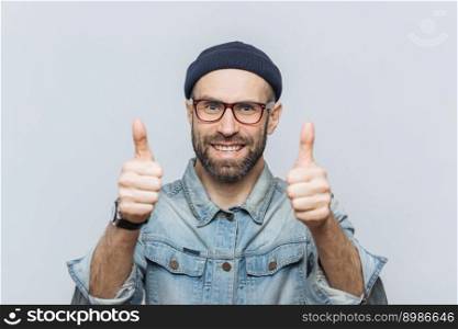 Happy bearded male model wears glasses, stylish hat and denim jacket, keeps thumbs raised, demonstrates approval and agreement with something, has delighted expression, isolated over grey wall