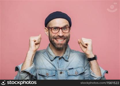 Happy bearded male with blue eyes and cheerful expression clenches fists, feels to be winner, demonstrates positiveness, isolated over pink background. Excited emotional young man poses indoor