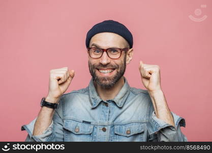 Happy bearded male with blue eyes and cheerful expression clenches fists, feels to be winner, demonstrates positiveness, isolated over pink background. Excited emotional young man poses indoor