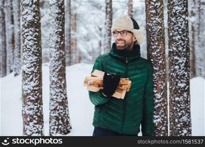 Happy bearded man in green jacket, stands near tree, holds firewood, looks thoughtfully aside, has cheerful expression, stands against snowy forest background. Fashionable pensive male outdoor