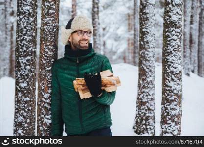 Happy bearded man in green jacket, stands near tree, holds firewood, looks thoughtfully aside, has cheerful expression, stands against snowy forest background. Fashionable pensive male outdoor
