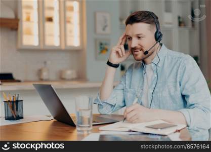 Happy bearded man with headset with microphone looking at laptop screen and making notes during distance education, sitting at his apartment while working online. Elearning and freelance concept. Smiling guy listening attentively online lesson while sitting at home
