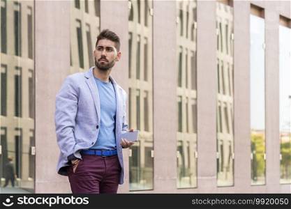 Happy bearded young man holding a smartphone while standing outdoors and looking away