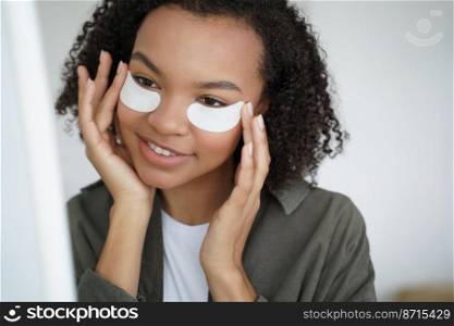 Happy beautiful african american girl applying moisturizing cotton under eye patches to reduce puffiness, remove dark circles. Beauty treatment for young skin, skincare cosmetics advertising.. Happy pretty african american young girl applying moisturizing under eye patches. Skincare routine