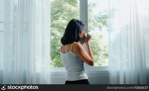 Happy beautiful Asian woman smiling and drinking a cup of coffee or tea near the window in bedroom. Young latin girl open curtains and relax in morning. Lifestyle lady at home concept.