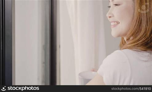 Happy beautiful Asian woman smiling and drinking a cup of coffee or tea near the window in the bedroom. Young asia female open the curtains and relax in the morning. Lifestyle woman at home concept.