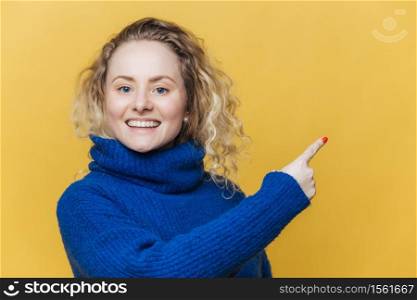 Happy beautiful blonde young woman has pleased expression, wears casual blue sweater, indicates at blank copy space on yellow blank studio wall, advertizes something with great pleasure. Advertisment