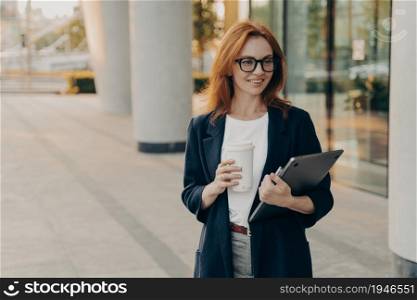Happy beautiful european red-haired businesswoman carrying laptop computer and cup of coffee on her way to office, female entrepreneur walking outdoors in urban city center. Stylish freelance people. Happy beautiful european red-haired businesswoman carrying laptop computer and cup of coffee
