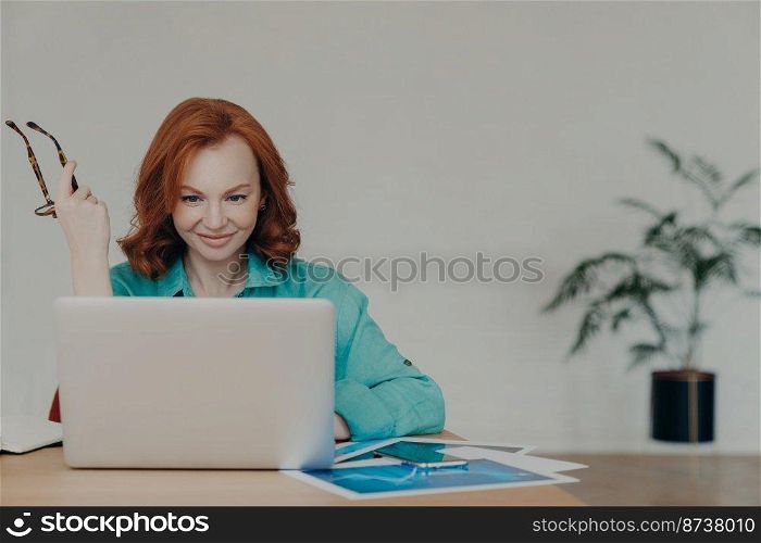 Happy beautiful female financial analyst prepares economic research, works on laptop computer, consults clients online, poses at desktop with documents, holds spectacles, busy with finishing task
