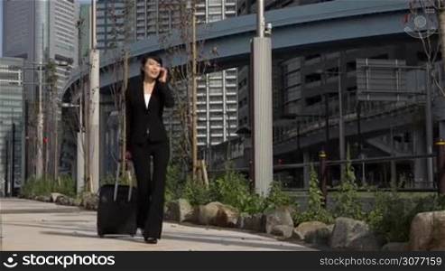 Happy beautiful Japanese female manager speaking on cell phone. Asian businesswoman, confident girl, busy woman talking with mobile telephone and walking with luggage during business trip. Slow motion