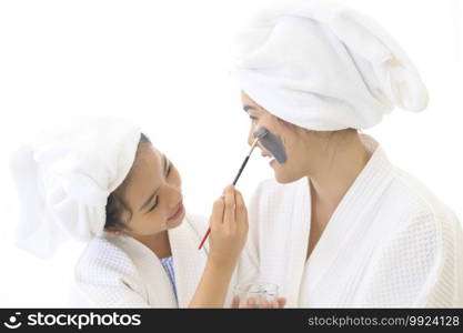 Happy beautiful mom and daughter in white bathrobe applying face mask in bedroom, Family and Beauty Concept. 