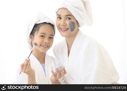 Happy beautiful mom and daughter in white bathrobe applying face mask in bedroom, Family and Beauty Concept. 