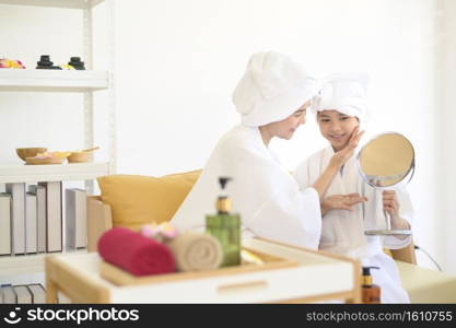 Happy beautiful mom and daughter in white bathrobe applying moisturizing cream on face at home, skin care and treatment concept

