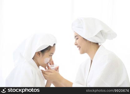 Happy beautiful mom and daughter in white bathrobe applying moisturizing cream on face at home, skin care and treatment concept  