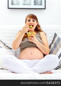 Happy beautiful pregnant woman sitting on sofa at home and eating fruit salad&#xA;