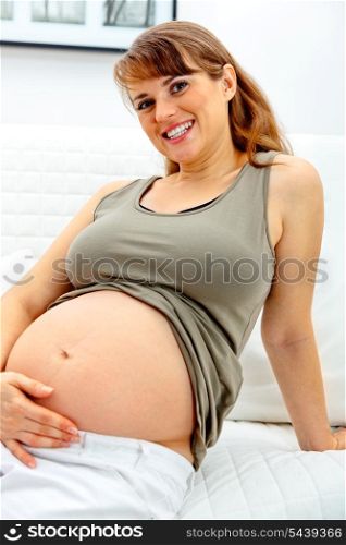Happy beautiful pregnant woman sitting on sofa at home and touching her belly.&#xA;