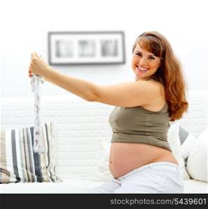 Happy beautiful pregnant woman sitting on sofa at home with baby clothes in hands.&#xA;
