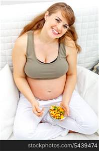Happy beautiful pregnant woman sitting on sofa at home with fruit salad in hand&#xA;