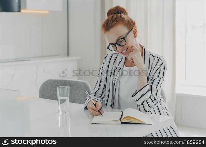 Happy beautiful redhead European woman makes notes in personal planner smiles gently holds pen poses at desktop drinks fresh pure water poses in spacious living room writes memo in her diary.. European woman makes notes in personal planner smiles gently holds pen poses at desktop
